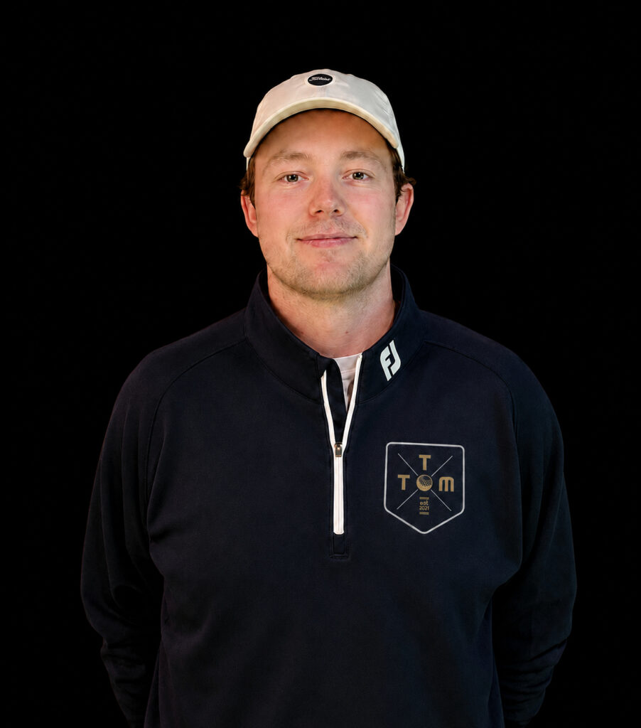 Portrait of a man in a navy pullover with a white zipper and a TOM logo on the chest, wearing a light-colored cap against a black backdrop.